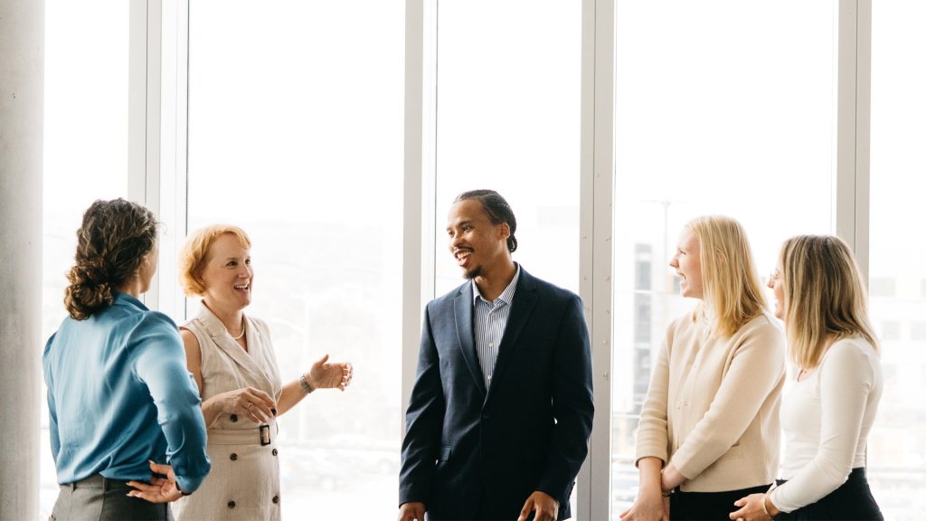 A group of five people standing near a large window engaged in a lively conversation. Everyone is smiling and interacting warmly.