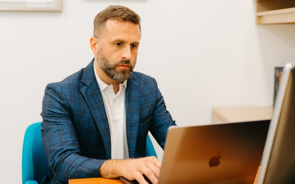 A man in a blue plaid blazer sits at a desk, focused on typing on a MacBook. The office setting includes a wooden bookshelf and clean, modern decor.