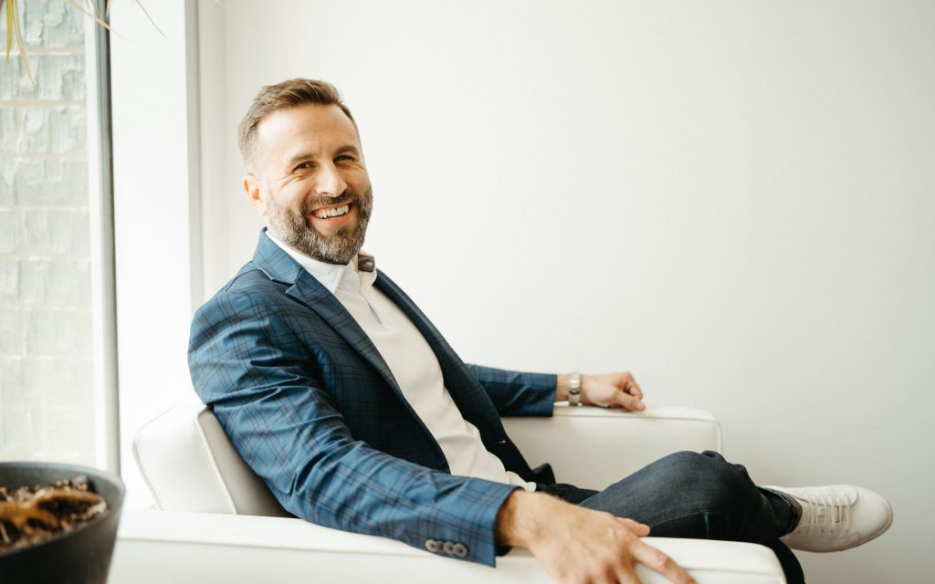 A man in a blue plaid blazer sits on a white chair, smiling warmly at the camera. He appears relaxed, with a bright, modern interior behind him.