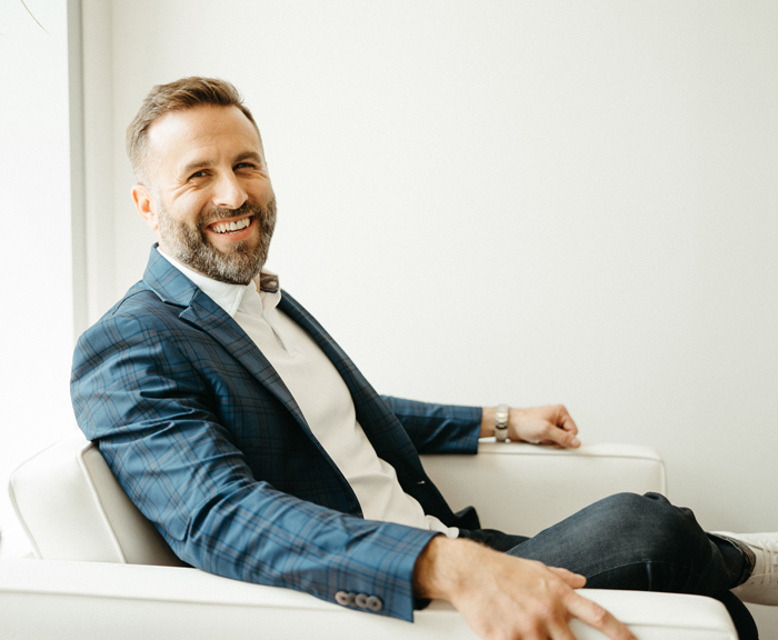 A man in a blue plaid blazer sits on a white chair, smiling warmly at the camera. He appears relaxed, with a bright, modern interior behind him.