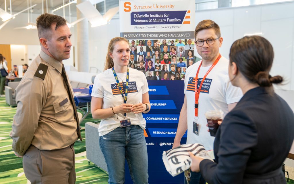 A male student in military uniform engages in conversation with two students wearing "Military and Veterans Law Society" T-shirts. Behind them is a Syracuse University D'Aniello Institute for Veterans & Military Families banner.