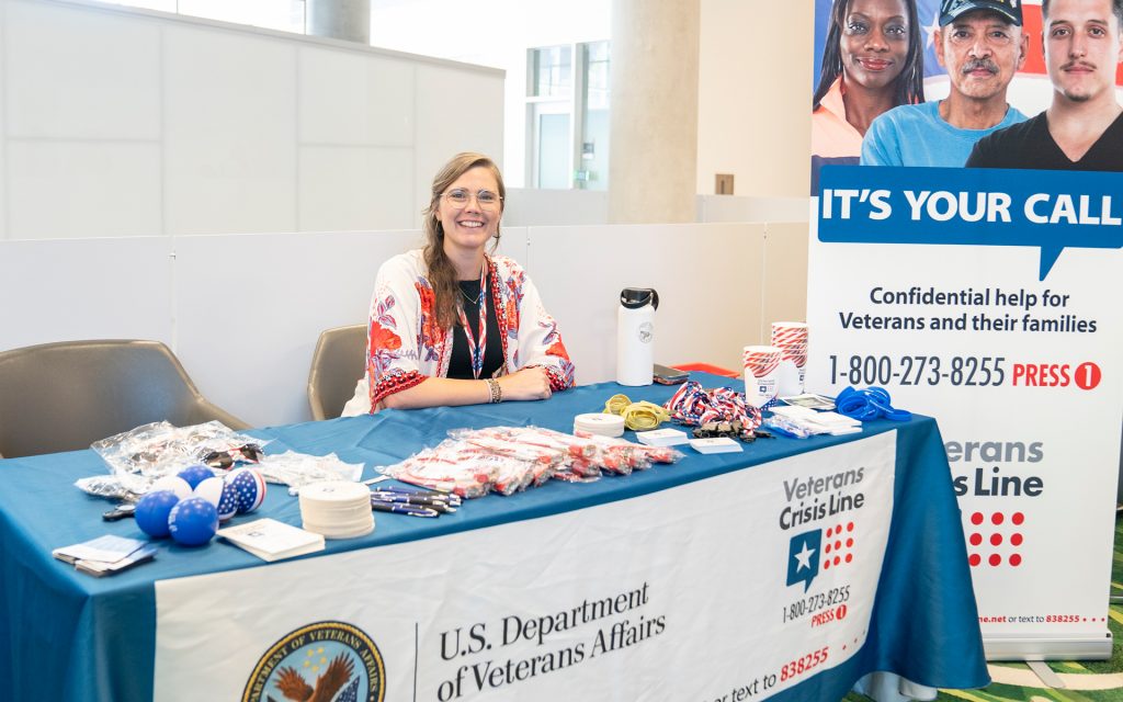 A female representative, seated at a table covered with a blue cloth, smiles at the camera. She sits beside promotional materials for the U.S. Department of Veterans Affairs and Veterans Crisis Line. A banner offers confidential help for veterans.