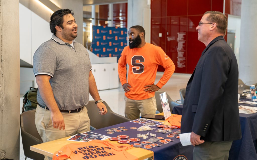  A group of three individuals converse near a table displaying "Syracuse University" and veteran-related promotional items. Two people stand, while one is seated behind the table.