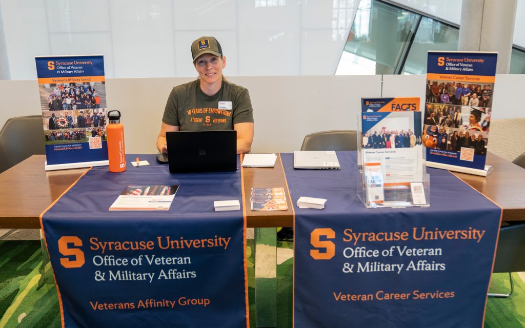 A Syracuse University representative sits at a table with banners promoting the Veterans Affinity Group and Veteran Career Services. She is wearing a cap and T-shirt, smiling at the camera.