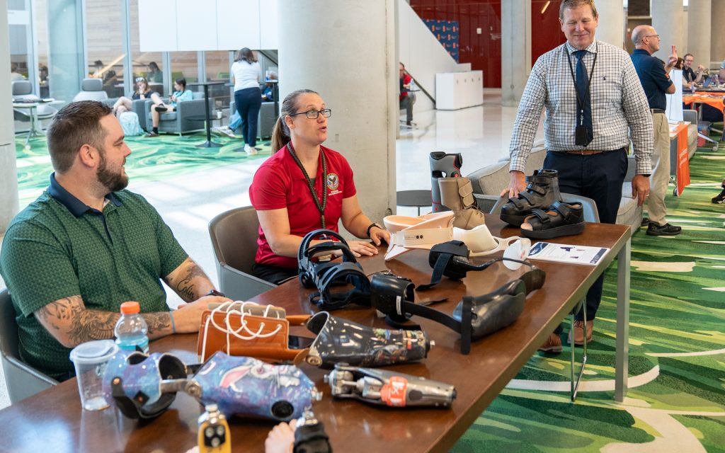 A group of individuals are seated at a table displaying prosthetic devices and orthotics for veterans. A man and woman explain the devices to a visitor standing by.