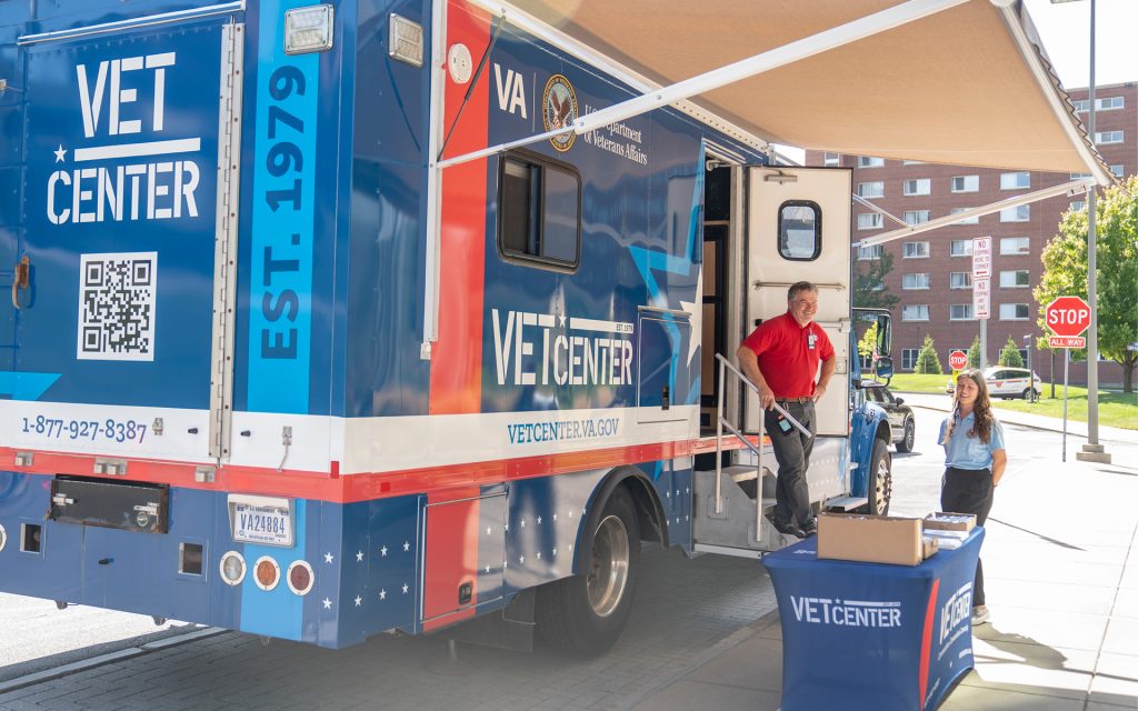 A large VA Vet Center truck parked outside, with two representatives from the Department of Veterans Affairs standing beside it. A banner and table with brochures are set up in front of the truck.