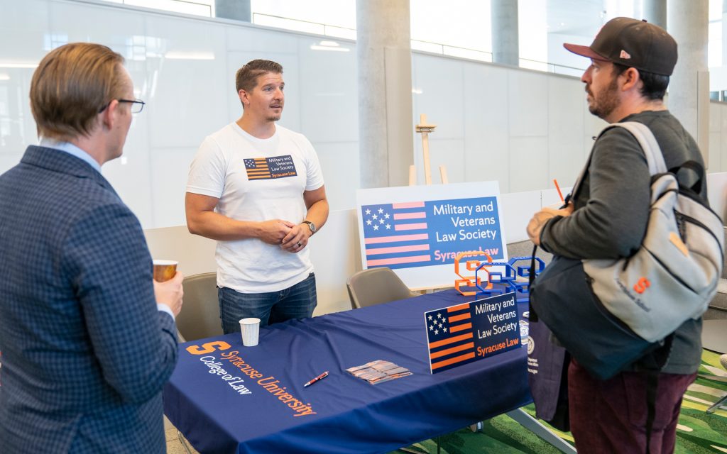 A Syracuse University Law student wearing a "Military and Veterans Law Society" T-shirt speaks with two other attendees near a table covered with promotional materials. A signboard promoting the society stands next to him.