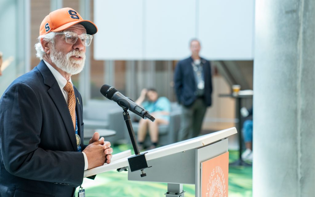 A man in a suit and an orange Syracuse University hat stands at a podium, speaking into a microphone. He is addressing attendees at the event inside a bright space with green flooring.