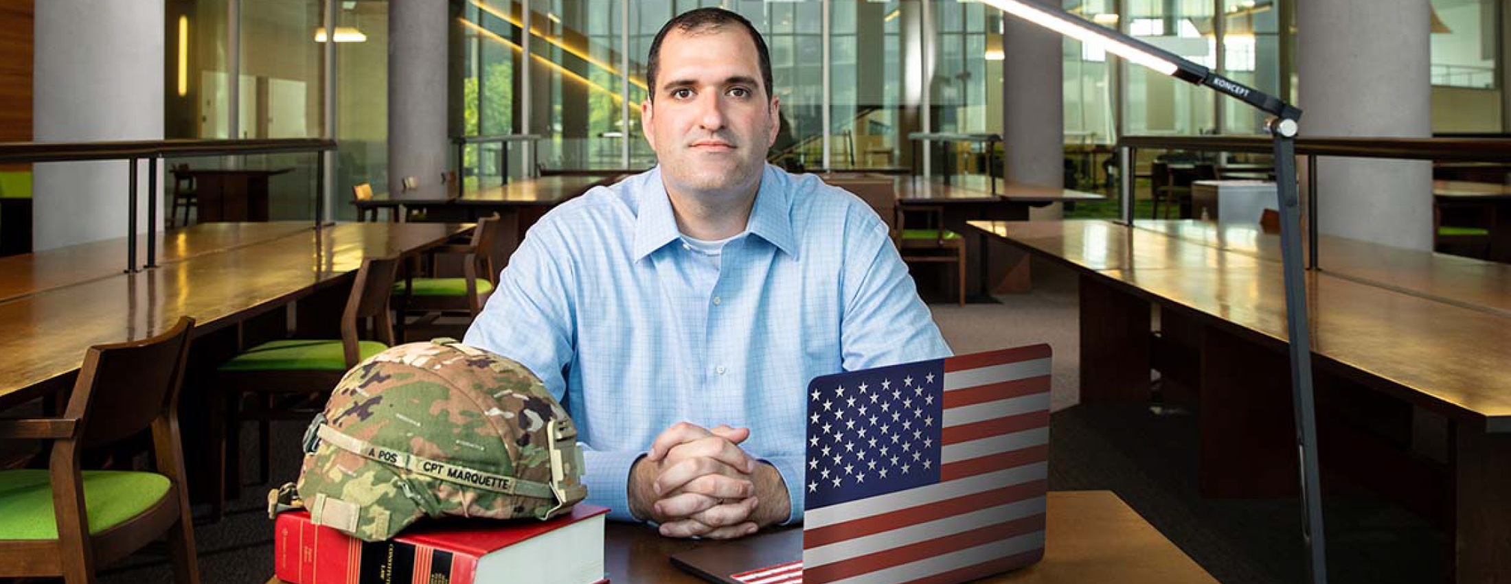 veteran sitting at a table in a library with his arms crossed, behind his army hat, a law book, and a laptop with a flag cover.