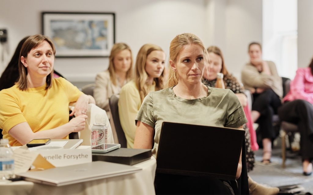 A group of attendees seated at a conference table during a presentation. In focus is a woman with long blonde hair, holding a laptop, appearing engaged with the speaker. Another woman in a yellow top sits nearby, along with other participants, in a well-lit room with framed art on the walls.