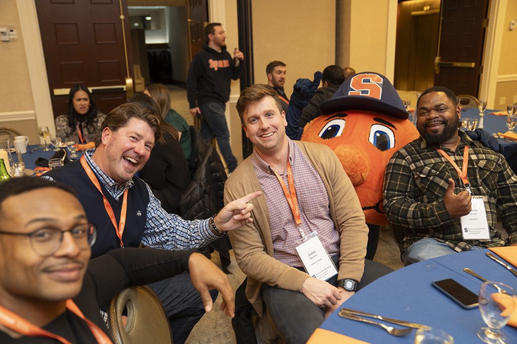 four students sitting around a table smiling, with otto the mascot in the background