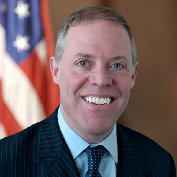 will barclay smiling at the camera in a black suit in front of the american flag