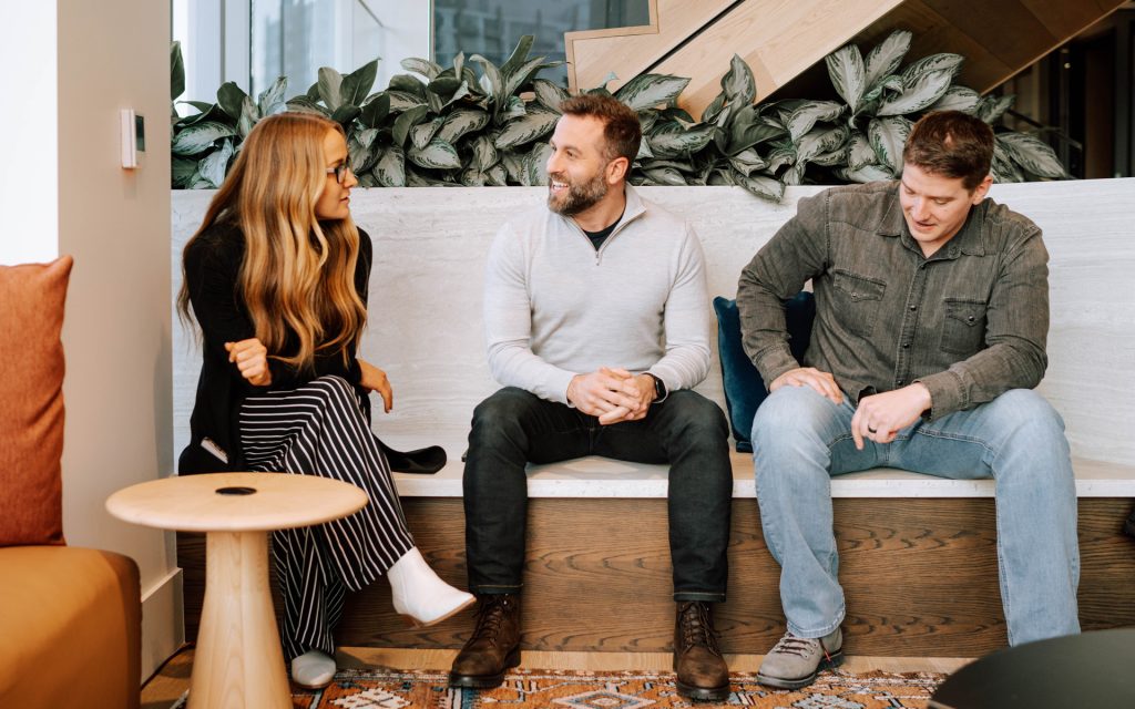 hree individuals sit together on a wooden bench, engaged in conversation. A woman with long blonde hair gestures while speaking to two men, one wearing a gray sweater and the other in a casual shirt and jeans. The setting includes modern furniture and greenery.