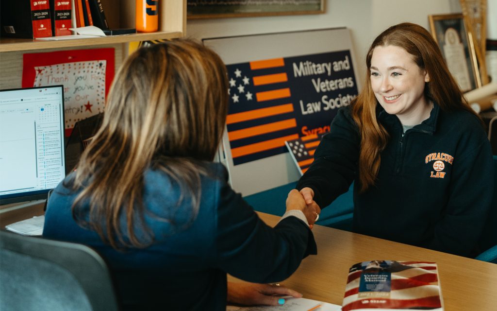 Meghan Wright shaking a woman's hand sitting at a desk in front of a Military and Veterans Law Society sign.