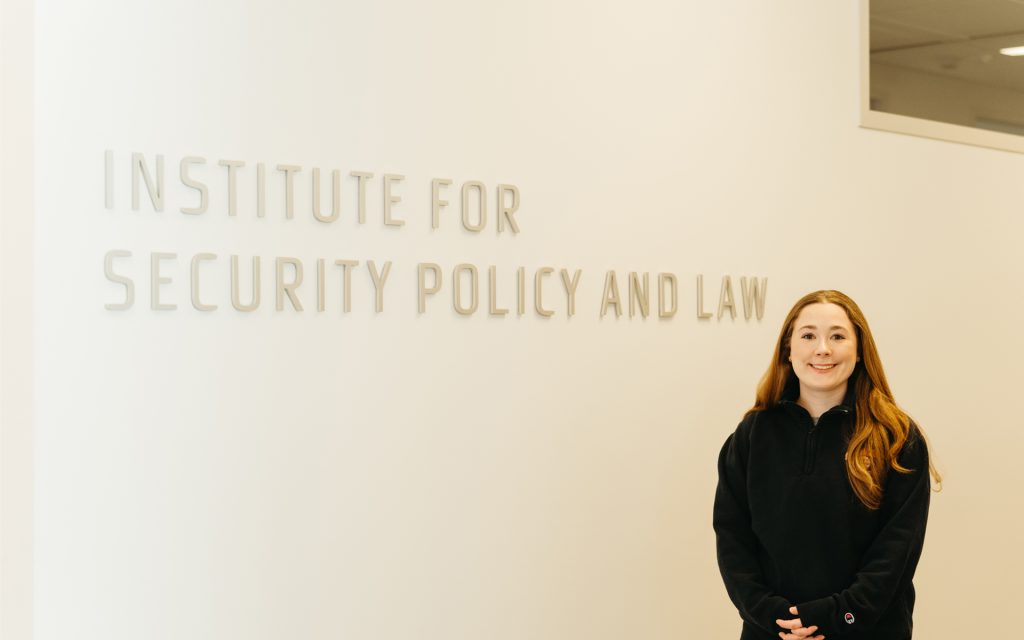 Meghan Write stands in front of a white wall and sign for the Institute for Security Policy and Law