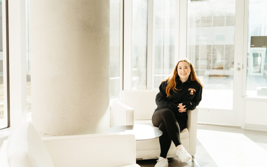 Meghan Wright sits in a white chair in the hallway of Dineen Hall in front of windows.