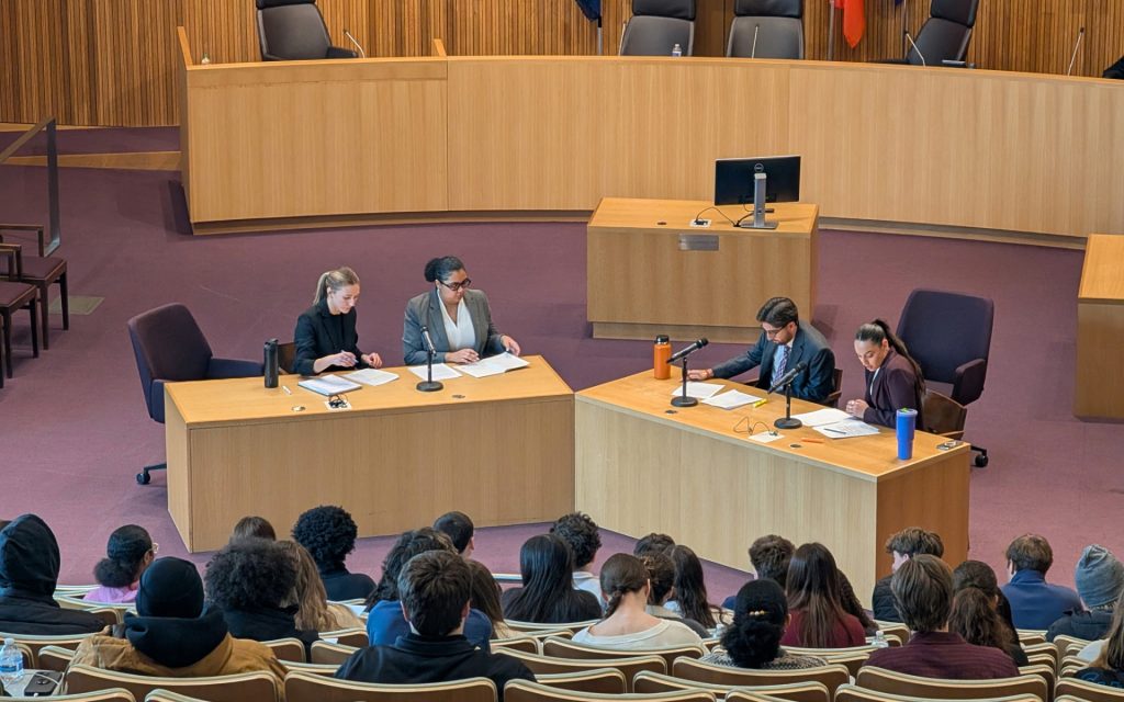 Four SU College of Law students in front of courtroom participating in an activity while students sit in the audience watching.