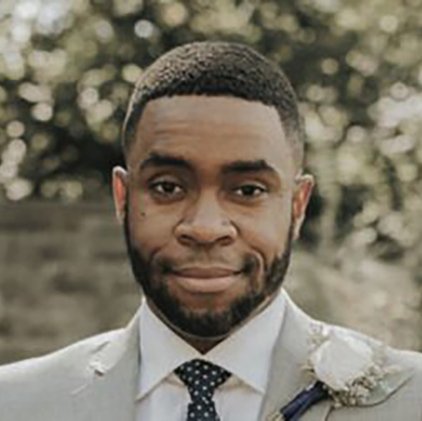 adom cooper grinning at the camera in a gray suit with a flower on his lapel in front of green shrubbery