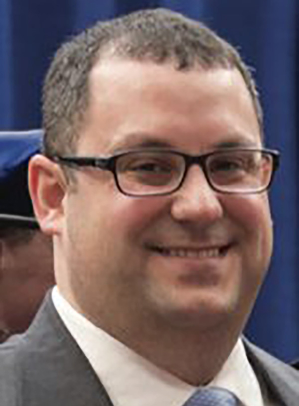 brian bauersfeld smiling at the camera in his headshot with black glasses on and a gray suit
