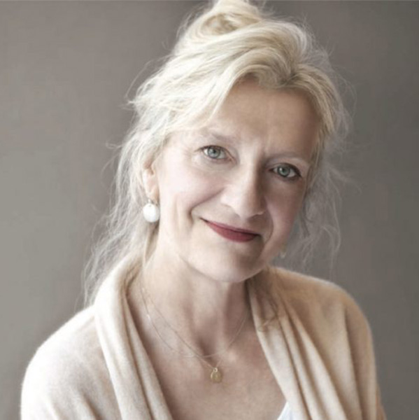 elizabeth strout grinning at the camera with a white cardigan on in front of a plain gray wall