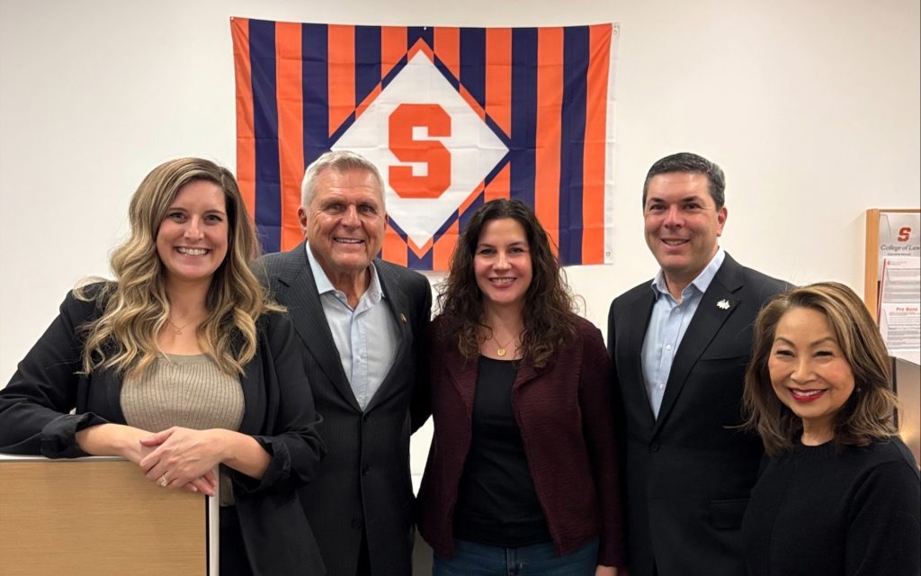 Pictured from left to right Adrienne Lutz, Michael Marino L’ 74, Dafni Kiritsis , Mike Marino L’96, Lily Hughes. Standing in front of Syracuse University flag.