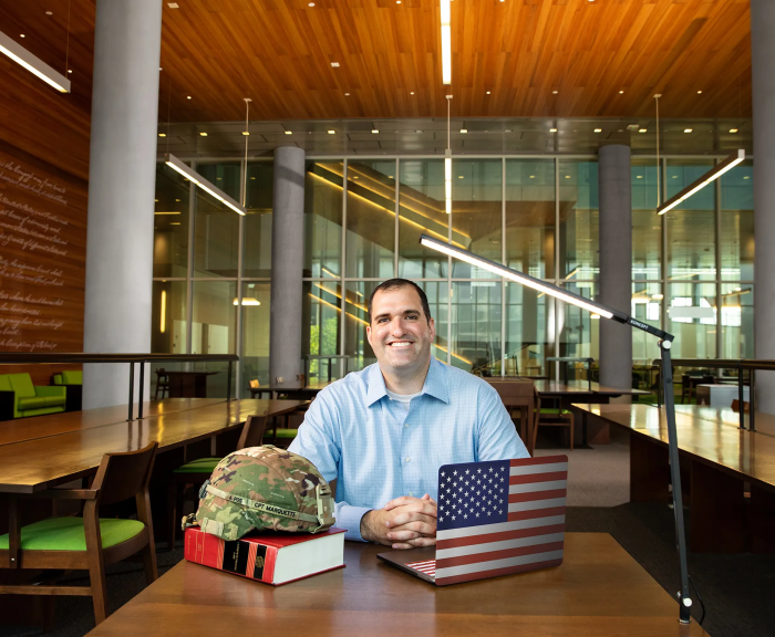 Ryan Marquette L'22 sitting in SU Law library with laptop with flag case, a large book and his military helmet on top. 