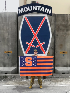 Ryan Marquette L’22 standing in military uniform with Syracuse University flag